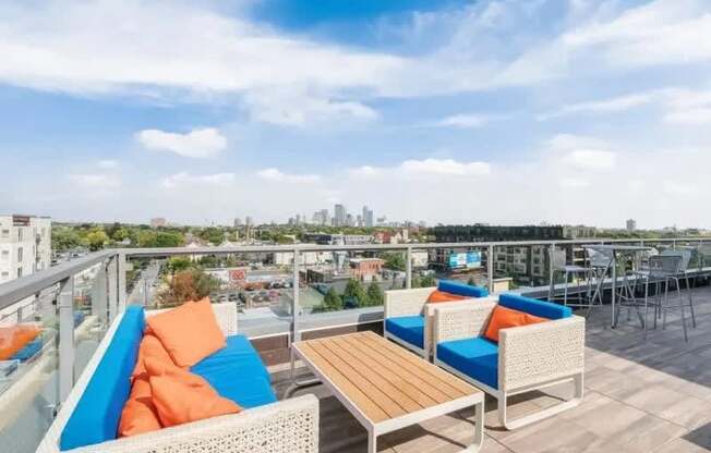 A stylish rooftop lounge here at Blue and Lime featuring woven outdoor seating with blue and orange cushions, a wood-top coffee table, and bar-height seating along a glass railing. Elevated views of the surrounding neighborhood and distant skyline create a relaxing setting for socializing, lounging, and enjoying open-air city scenery.