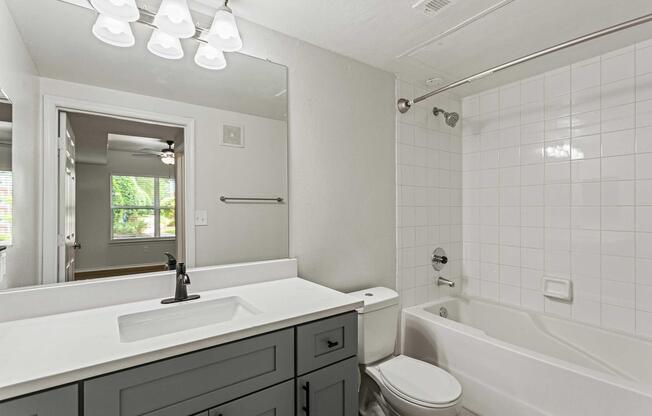 Modern bathroom featuring a white tiled wall, a bathtub with a shower, and a sleek gray vanity. The sink area has a black faucet, and overhead lights illuminate the space. A door leads to a room with a ceiling fan and window, adding natural light to the overall design.