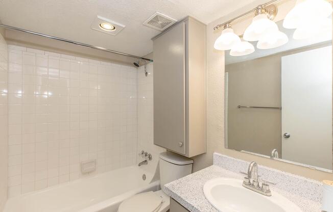 A clean, modern bathroom featuring a white tiled shower and bathtub combination, a toilet, and a granite countertop with a sink. The space is well-lit by three frosted glass light fixtures above the mirror, and there is a neutral color scheme throughout.