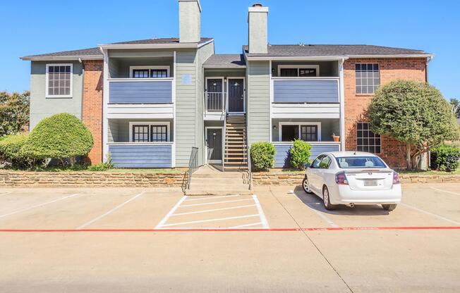 A two-story apartment building with a combination of brick and gray siding. There are balconies on the upper level and a set of stairs leading to the entrance. The parking lot in front has a white car parked next to the building, with neatly trimmed bushes and a clear sky in the background.