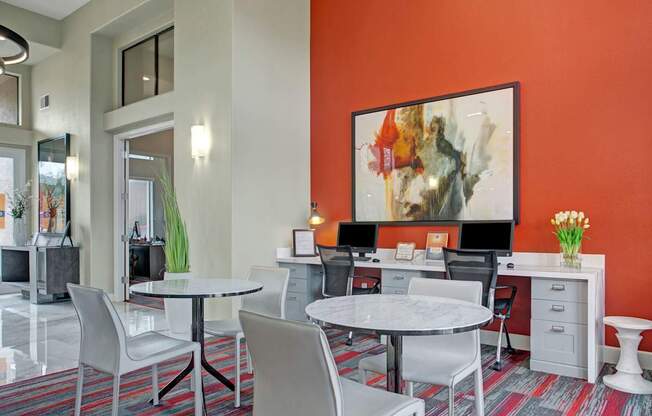 A modern business center here at Mirador featuring a bold red accent wall with large abstract artwork, built-in workstations with computers and ergonomic chairs, and sleek marble-top tables for collaborative seating. Neutral tones and contemporary lighting create a bright, professional space ideal for remote work or study.