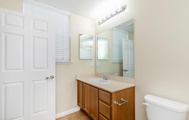 A clean and modern bathroom featuring a light-colored wall, a single sink with wooden cabinet storage, two mirrors above the sink, and a white toilet. Natural light filters in through a window with blinds, and a door is partially open, revealing another room. The floor is tiled in a neutral hue.