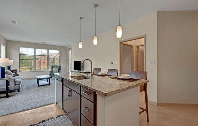 A bright open kitchen and living area here at Vue at Pinnacle Park with a granite island, bar seating, pendant lighting, and stainless sink, flowing into a spacious carpeted lounge with large windows and natural light.