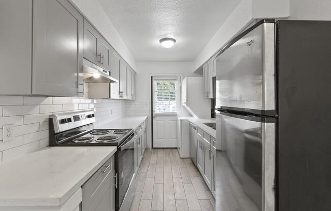 A modern kitchen featuring gray cabinetry, stainless steel appliances, and a white tile backsplash. The kitchen includes an electric stove, refrigerator, and ample counter space. A door leads to an outdoor area, and a window allows natural light to fill the space. Light wood-look flooring adds a warm touch.