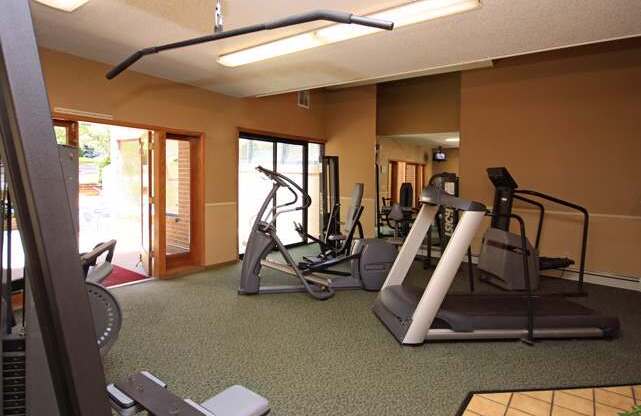 A spacious fitness center here at Eagle Ridge featuring multiple cardio machines including treadmills and an elliptical, along with strength training equipment arranged on green carpeted flooring, with overhead lighting, neutral walls, and large windows and glass doors that bring in natural light and views of the surrounding community.