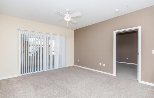 A spacious living area featuring a ceiling fan, light-colored walls, and carpet flooring. One wall has sliding glass doors leading to an outdoor area, while another wall has an open doorway to an adjacent room. The space appears well-lit and inviting, with a neutral color palette.