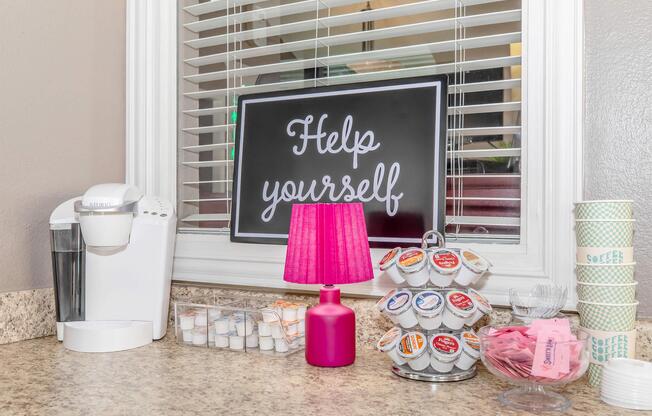 A cozy coffee corner featuring a black sign that reads "Help yourself," a pink lamp, a coffee maker, a carousel of coffee pods, and colorful cups on a countertop near a window with blinds. There are also a few small containers with sugar or sweeteners.