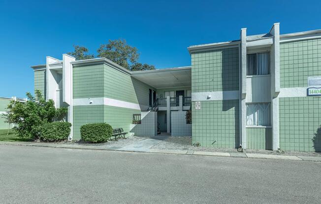 A modern two-story apartment building with green and white exterior. The entrance features a small set of stairs leading to the door, flanked by low-maintenance landscaping. Clear blue skies in the background. The building has a clean, minimalist design with large windows.
