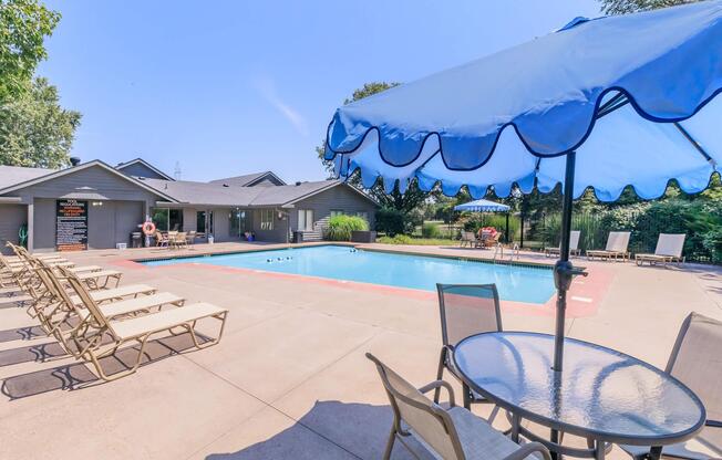 A swimming pool surrounded by lounge chairs and tables. A blue umbrella provides shade over a table. In the background, there are several houses and lush greenery. The sky is clear and blue, indicating a sunny day. The pool area offers a relaxing outdoor space for leisure.
