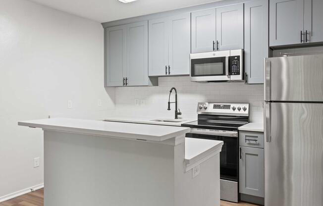 Modern kitchen featuring light gray cabinets, stainless steel appliances including a microwave and stove, a sleek countertop, and a minimalist design. Natural light enhances the space, which has a clean, contemporary look with wooden flooring.