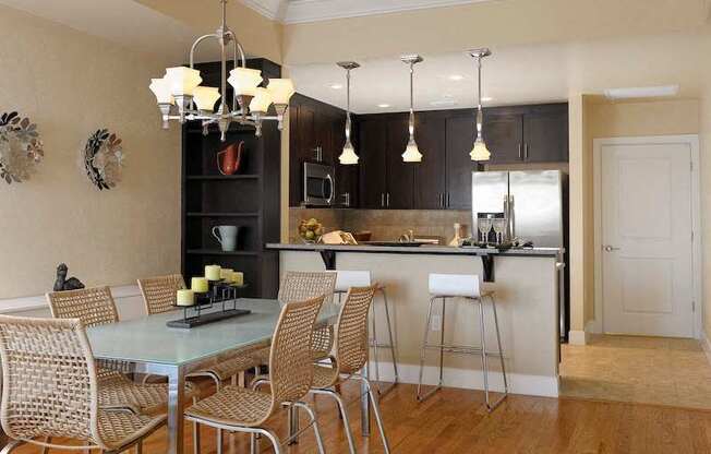 A modern kitchen with stainless steel appliances, hardwood-style flooring, pendant lights, a breakfast bar, and a built-in corner shelving unit near a dining area.