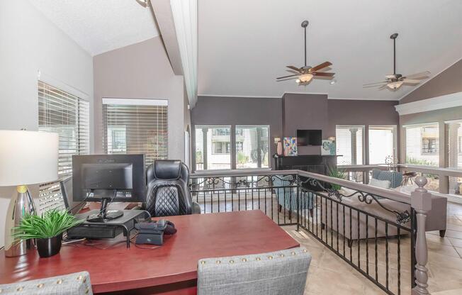 Modern interior of a home office featuring a wooden desk with a computer, a comfortable black office chair, and a decorative plant. The room has large windows allowing natural light, ceiling fans, and a stylish balcony railing. Cozy seating areas are visible in the background, creating an inviting atmosphere.
