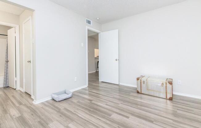 A lightly furnished room with light gray walls and new laminate flooring. There is a closed door on the left, an open door leading to another area, and a decorative trunk in the center. A small pet bed is positioned on the floor. The space appears clean and minimalistic, with ample natural light.