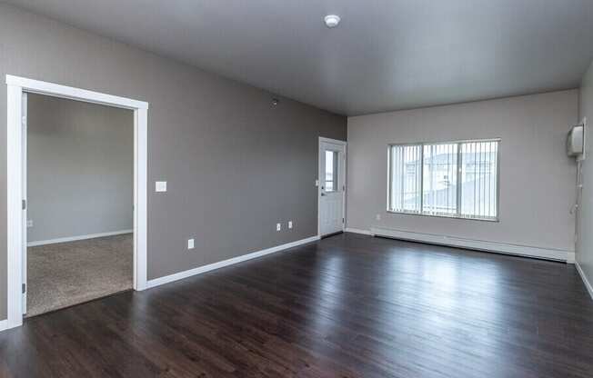 A large empty room with wood flooring and a window with blinds.