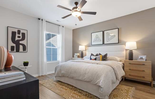 A primary bedroom with an illuminated ceiling fan, large window, model furnishings, and wood-style flooring