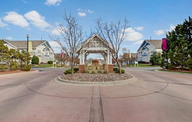 A charming community entrance here at Arbor Ridge featuring a decorative gazebo, landscaped roundabout, and welcoming residential buildings. Mature trees and colorful banners create a warm, inviting neighborhood setting.