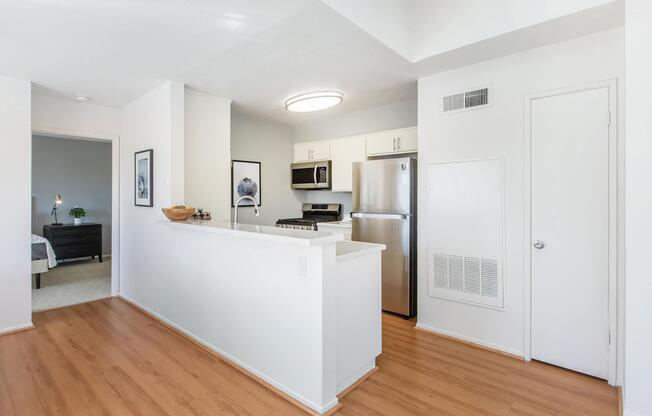 Bright, modern kitchen with white cabinetry and stainless steel appliances, including a refrigerator and stove. Light wood flooring extends into a living space, with an open layout. A doorway leads to a softly lit bedroom area in the background. The overall ambiance is clean and inviting.