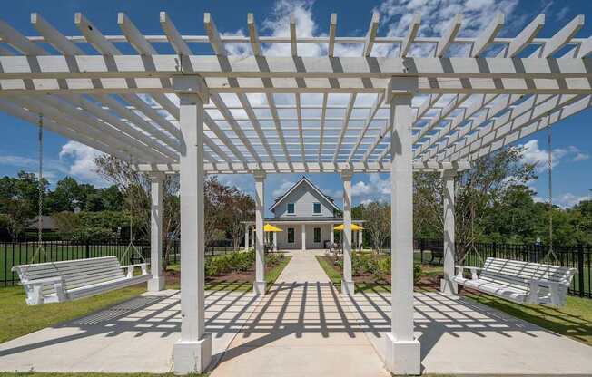 A white pergola structure is in the foreground of a park with benches and a building in the background.