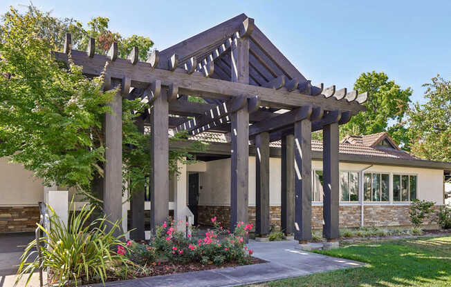 A wooden pergola with a walkway and flowering plants in front of a house.