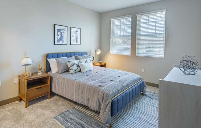Studio Apartments In Burlingame, CA - Bayswater Burlingame - Spacious Bedroom With Plush Carpet Flooring, Area Rug, Bed, Nightstands With Lamps, Dresser, Wall Art, And Two Windows With Blinds at Baysw