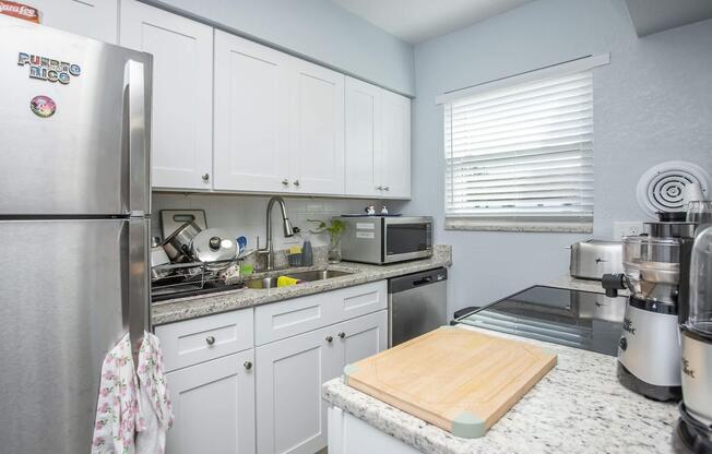 A modern kitchen featuring white cabinetry, a granite countertop, a stainless steel refrigerator, a microwave, and a stove. There are dishes in a drying rack near the sink. A wooden cutting board rests on the counter, and light streams in through a window with white blinds.