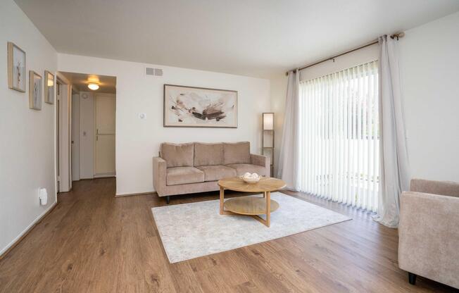 A cozy living room featuring a light brown sofa, a round coffee table with a decorative bowl, and a large window covered with vertical blinds. The room is adorned with framed artwork on the walls, soft lighting, and a neutral color palette, creating a warm and inviting atmosphere.