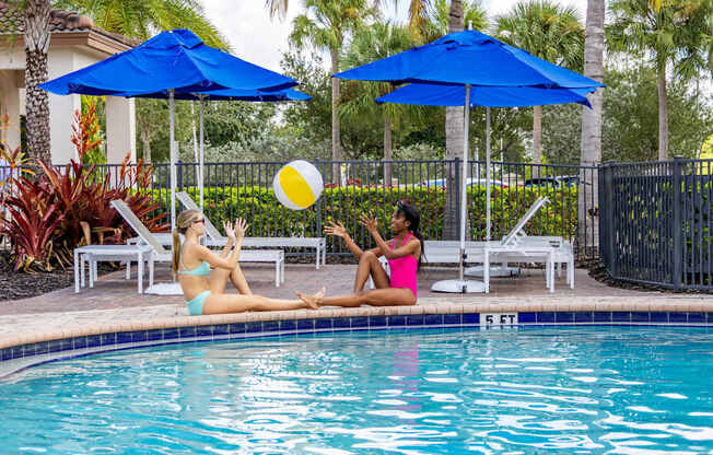 Two women sunbathing by a pool with blue umbrellas.