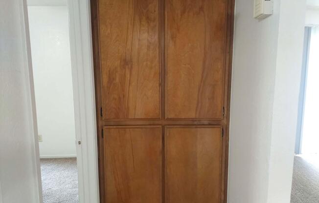 Wooden cabinet with four flat-panel doors, located in a hallway. The cabinet's rich brown finish contrasts with the light-colored walls and carpet. A white wall with a small thermostat is visible to the right, while the hallway extends into a brighter room beyond.