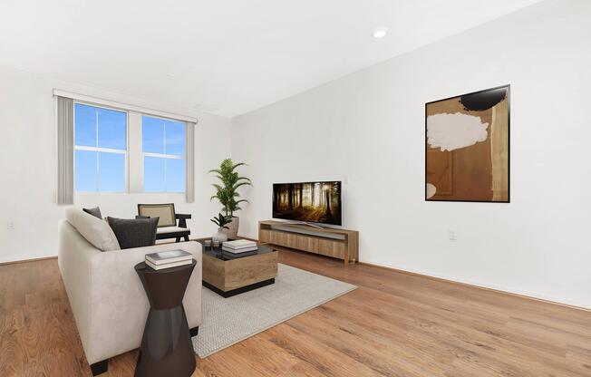 A modern living room featuring a light-colored sofa with black and white accent pillows, a wooden coffee table, and a flat-screen TV on a light wood media console. A decorative plant is in the corner, and a piece of abstract art is hanging on the wall. The room has hardwood flooring and large windows allowing natural light to enter.