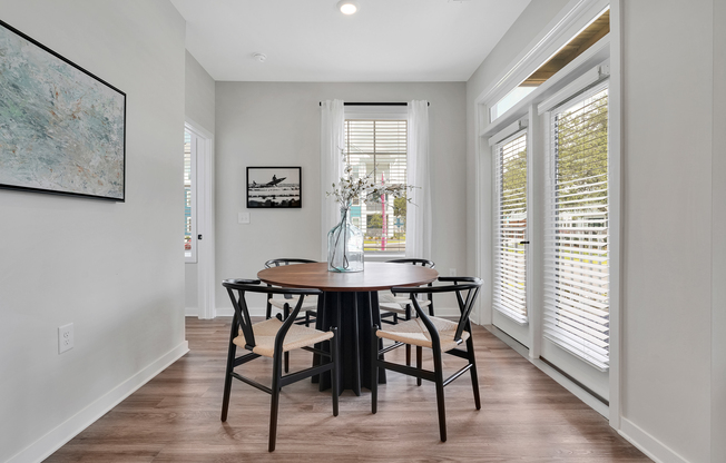 A modern dining area featuring a round wooden table surrounded by four black chairs. Large windows with white blinds allow natural light to fill the space. A vase with flowers sits on the table, and artwork hangs on the wall, adding to the cozy and inviting atmosphere.