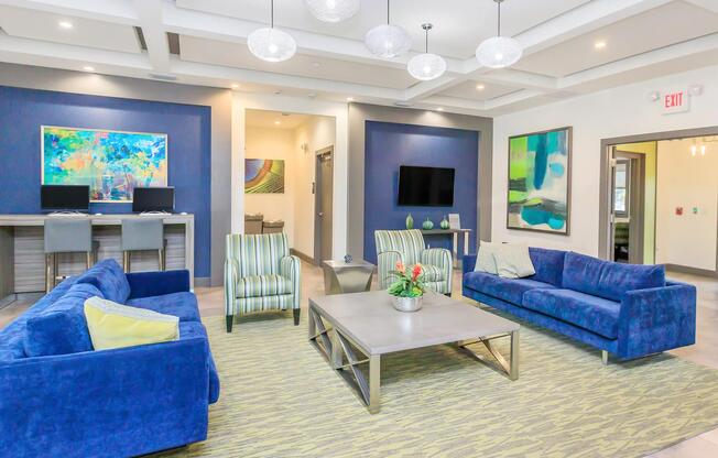 A modern lounge area featuring blue sofas arranged around a coffee table, vibrant artwork on the walls, striped chairs, a rug, and lighting fixtures. In the background, there are two computer workstations and a wall-mounted TV, creating a welcoming and stylish environment.