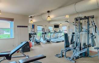 A spacious, well-lit fitness center here at Dakota Ridge featuring treadmills lined along large windows, a multifunction weight machine, a padded workout bench, neutral carpet flooring, exposed ceiling ductwork, and warm pendant lighting that creates a clean, motivating environment for residents to exercise comfortably.