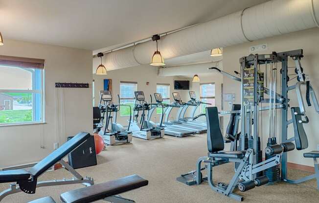 A spacious, well-lit fitness center here at Dakota Ridge featuring treadmills lined along large windows, a multifunction weight machine, a padded workout bench, neutral carpet flooring, exposed ceiling ductwork, and warm pendant lighting that creates a clean, motivating environment for residents to exercise comfortably.