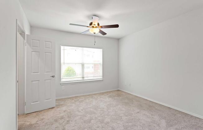 A light and airy bedroom with beige carpet, a ceiling fan, and a large window allowing natural light. The walls are painted in a neutral tone, and there is a doorway leading to another room. The space appears clean and minimalistic, suitable for various decor styles.