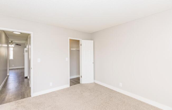 Empty room with light gray walls and carpeted floor. A closed white door on the right leads to a closet, while another door on the left opens to a hallway. The space is well-lit with a ceiling light fixture visible in the background, suggesting a clean and modern living environment.