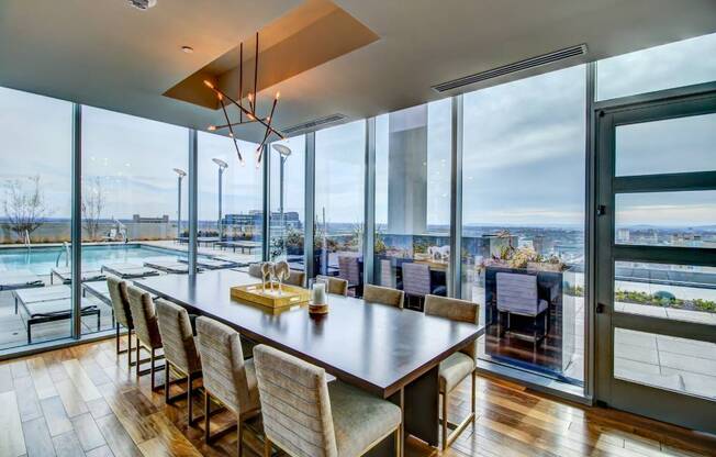 A modern dining room with a long wooden table and chairs, a chandelier, and a view of the outdoors through large windows.