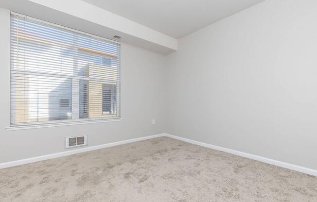 A spacious, empty room with light beige carpet and neutral-colored walls. A large window with vertical blinds allows natural light to enter, revealing a glimpse of an exterior wall and windows. The room has no furniture, creating a clean and open space.