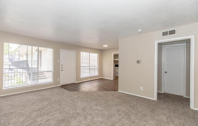 Spacious interior of a well-lit apartment featuring a carpeted living area with large windows. A door leads to a balcony. The room connects to a kitchen visible through an open doorway and includes a closet on the right, enhancing the apartment's functionality and inviting atmosphere.