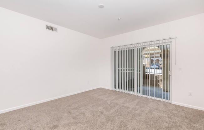Empty room with light-colored walls and carpet flooring. A large sliding glass door with vertical blinds opens to an outdoor space. The room features natural light and minimal decor, providing a spacious and airy ambiance.