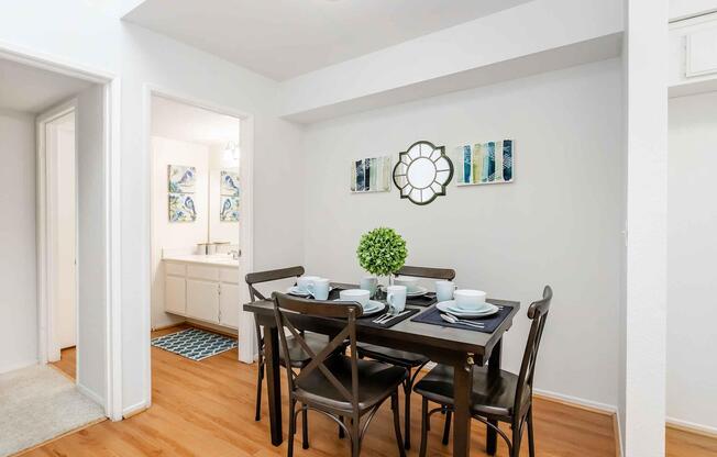 A cozy dining area featuring a dark wooden table set for four with plates and cups. A small green plant sits in the center of the table. The wall displays a decorative mirror and abstract artwork, while a doorway leads to a well-lit bathroom area. Light wooden flooring complements the bright, neutral walls.