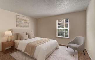 A bright bedroom here at The Bradford with beige carpet, soft neutral walls, and a large window with blinds allowing natural light to fill the space. A neatly staged bed with neutral bedding and a simple accent chair create a calm, comfortable, and inviting atmosphere.