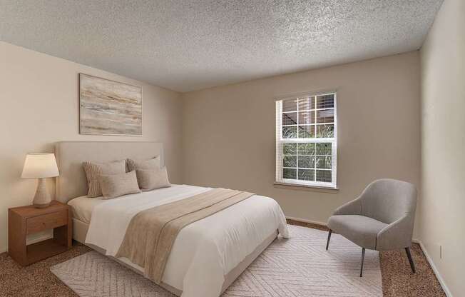 A bright bedroom here at The Bradford with beige carpet, soft neutral walls, and a large window with blinds allowing natural light to fill the space. A neatly staged bed with neutral bedding and a simple accent chair create a calm, comfortable, and inviting atmosphere.