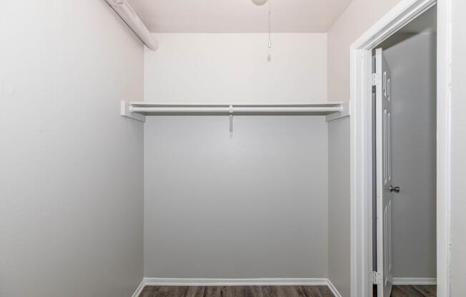Empty closet with a single hanging rod and light-colored walls. The floor is covered in a wooden laminate. A doorway on the right leads to another room. The space is unadorned, creating a minimalist look.