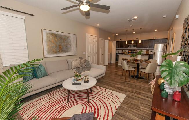 A modern living room featuring a light-colored sectional sofa, round coffee table, and decorative throw pillows. The space includes a dining area with a small table and chairs, a well-equipped kitchen in the background, and indoor plants for added decor. Soft lighting enhances the warm, inviting atmosphere.