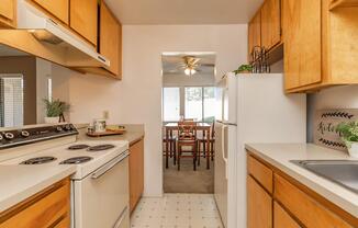 A view of a modern kitchen featuring wooden cabinets, a white stove, and refrigerator. An open doorway leads to a dining area with a table and chairs, illuminated by natural light coming through windows. The kitchen has a clean, functional layout with a sink and decorative touches.