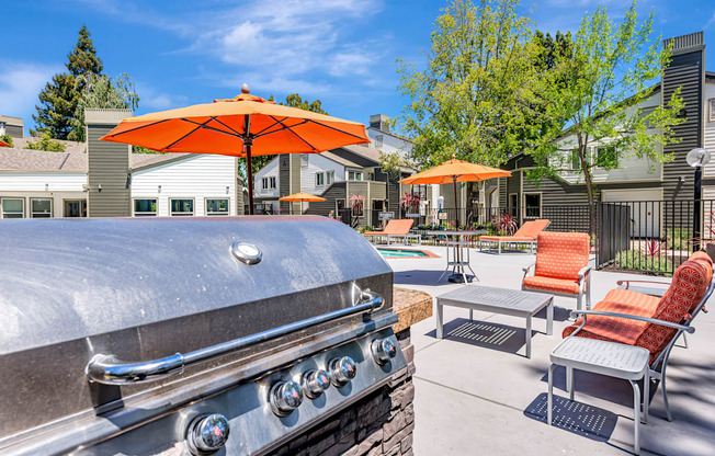A silver grill is in the foreground of a patio with orange chairs and umbrellas.