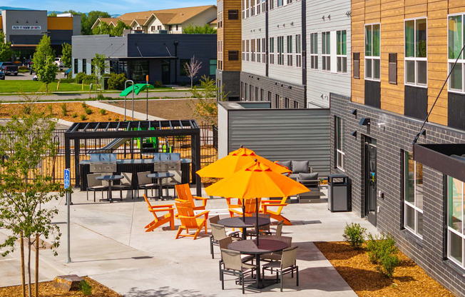 A patio with orange chairs and tables is surrounded by buildings.