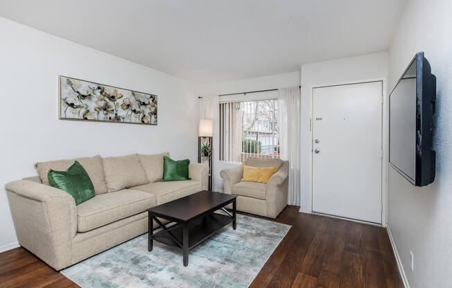 A cozy living room featuring a beige sofa with green accent pillows, an armchair, and a dark wood coffee table. A light-colored area rug lies on the floor, and a large abstract painting hangs on the wall. Natural light filters through sheer curtains at the window, and a front door is visible.