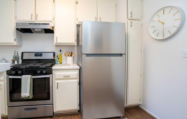 A modern kitchen featuring a silver refrigerator beside a black stove with an oven. The cabinets are light-colored, and a wall clock with a minimalist design is mounted nearby. The countertop is clean, adorned with utensils and a bottle, reflecting a tidy and functional cooking space.