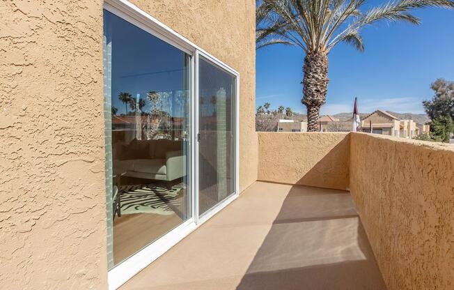 A corner balcony with a smooth beige floor and a concrete wall, featuring a large glass door that leads to an interior space. Outside, there are tall palm trees and a clear blue sky in the background, with residential buildings visible. The balcony offers natural light and a relaxing outdoor atmosphere.